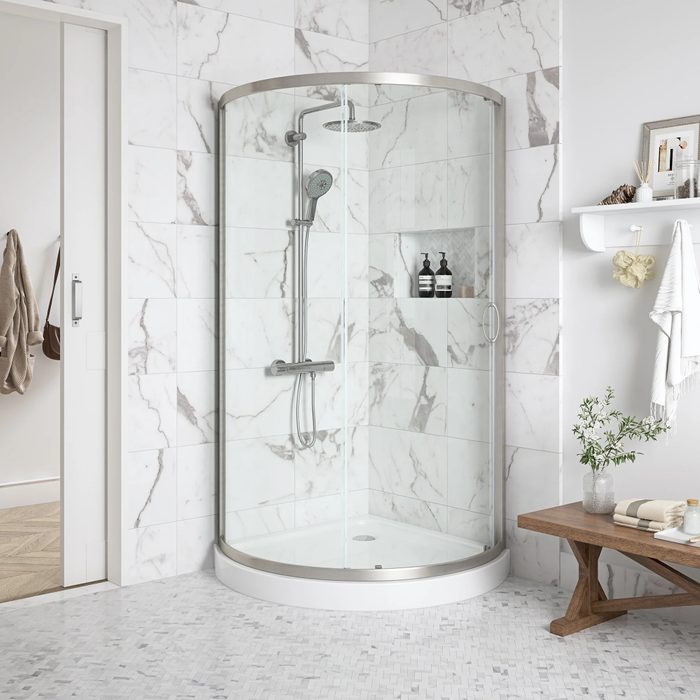 A glass shower enclosure features a showerhead and wall-mounted controls. It sits on marbled tile flooring, surrounded by light-colored walls and a wooden table.