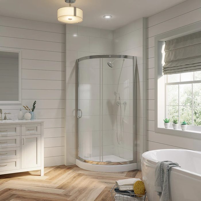 A glass shower stall stands in a modern bathroom, featuring a vanity and window with natural light illuminating the wooden floor and decorative plants.