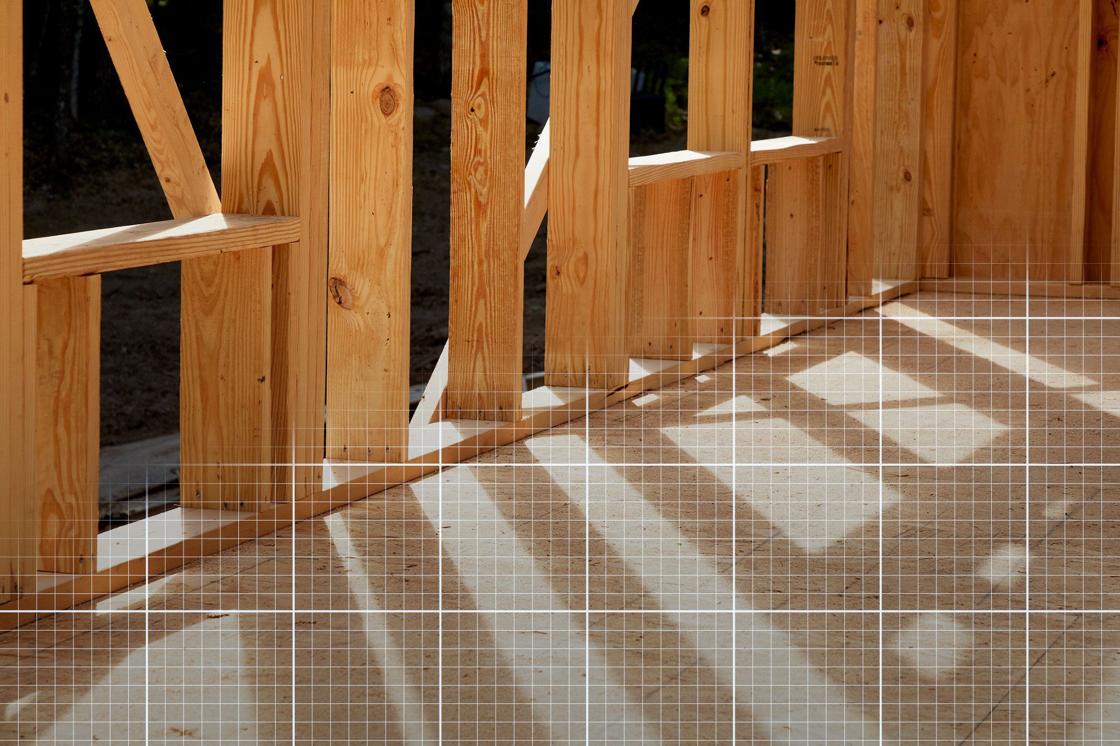 Wooden frame walls cast shadows on a light-colored floor, indicating ongoing construction in a bright, natural environment. Sunlight filters through open spaces.