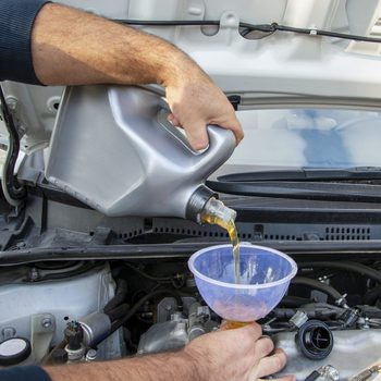 A hand pours engine oil from a container into a funnel, directing it into the engine compartment of a vehicle, under a raised hood.