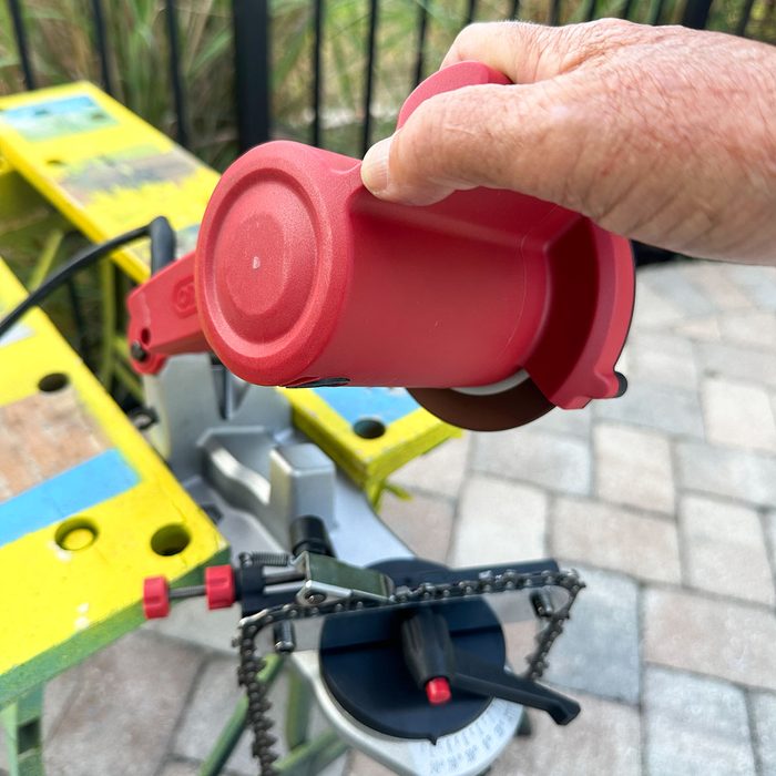 A hand holds a red tool against a circular saw, set on a multicolored wooden surface, surrounded by a blurred outdoor background.