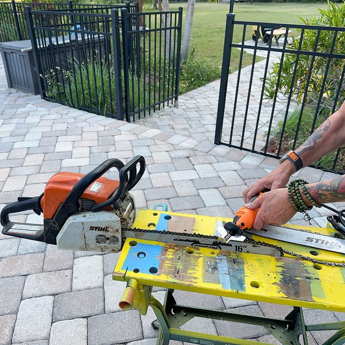 A chainsaw rests on a yellow workbench while a person adjusts its blade, surrounded by a paved outdoor area with grassy borders and a fence.