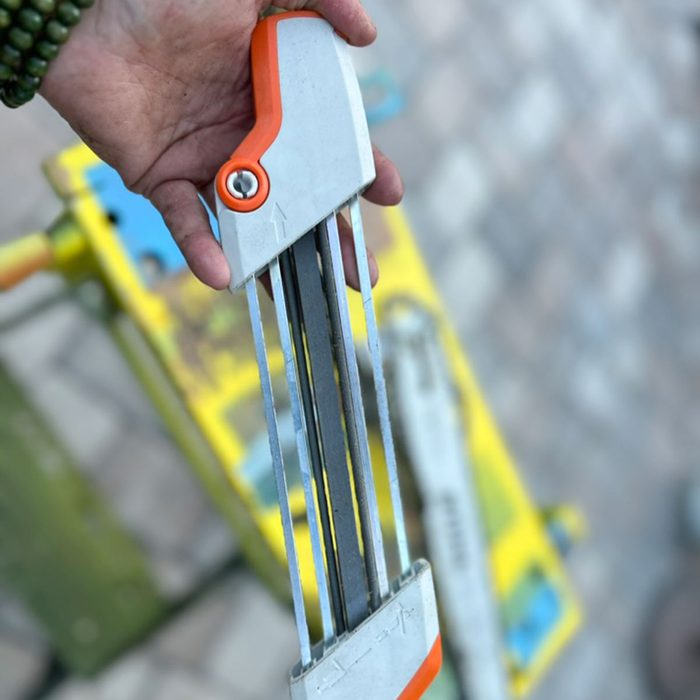 A hand holds a multi-blade tool, possibly for cutting or trimming, against a blurred background of a colorful work surface.