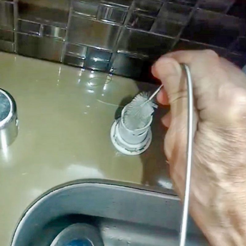 A hand grips a cleaning tool, scrubbing a drain in a shiny sink, surrounded by a tiled backsplash. Water glistens on the surface.