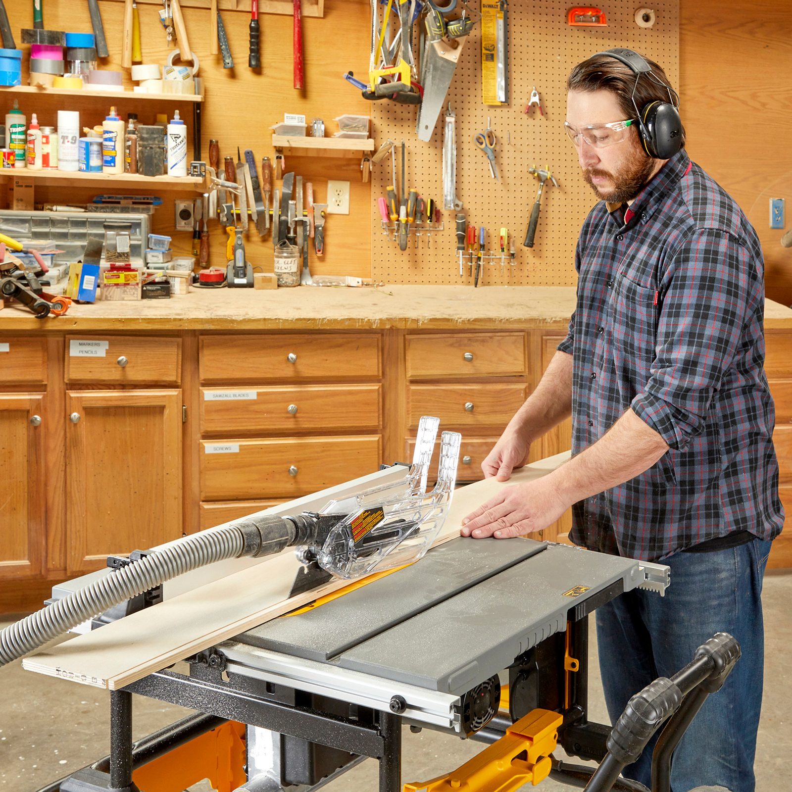 A man operates a table saw, guiding a piece of wood while wearing protective gear, surrounded by tools and materials in a workshop.