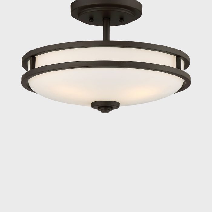 A ceiling light fixture illuminates a space, featuring a round white glass shade encased in dark metal rings against a plain background.