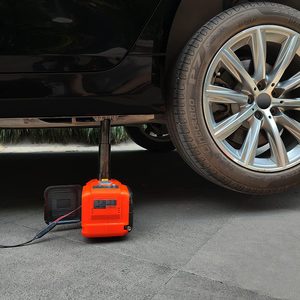 An orange hydraulic jack supports a car, lifting it off the ground. The vehicle is parked on a paved surface next to a shiny wheel.