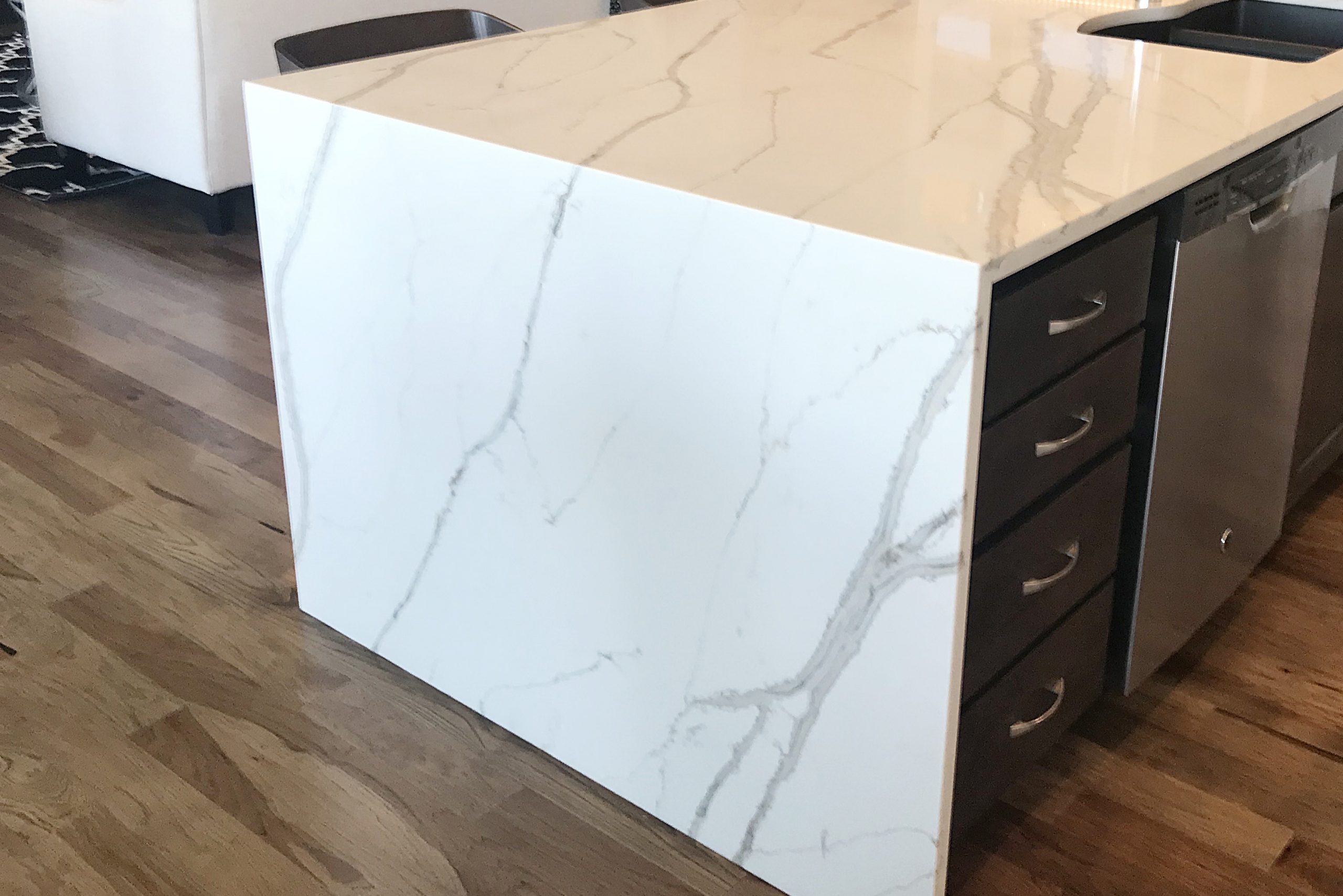 A modern kitchen island featuring a white stone surface with gray veining, integrated drawers, and adjacent stainless steel appliances, set on a wooden floor.