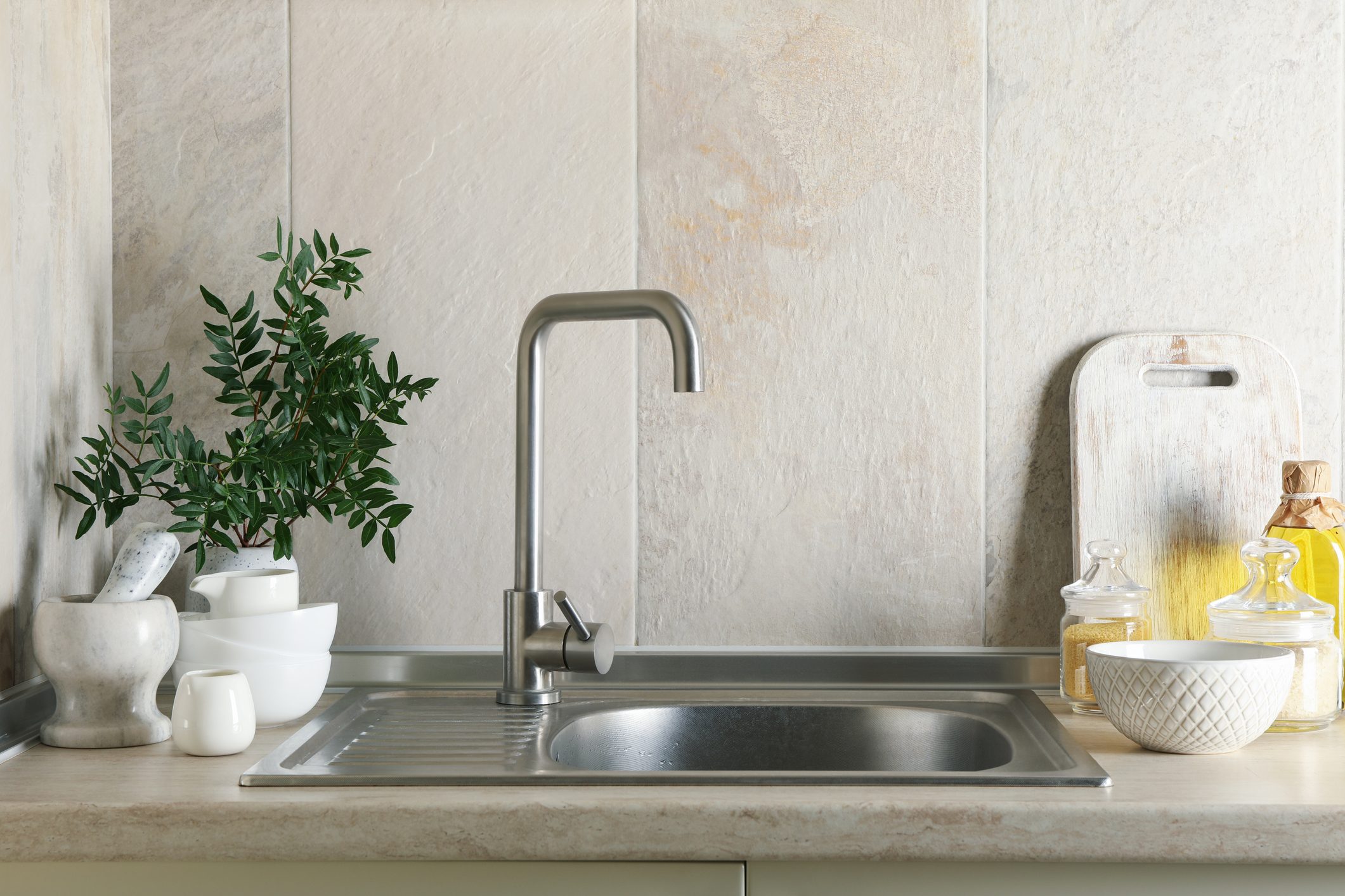 A stainless steel sink with a modern faucet sits on a beige countertop, surrounded by various kitchenware and a small green plant.