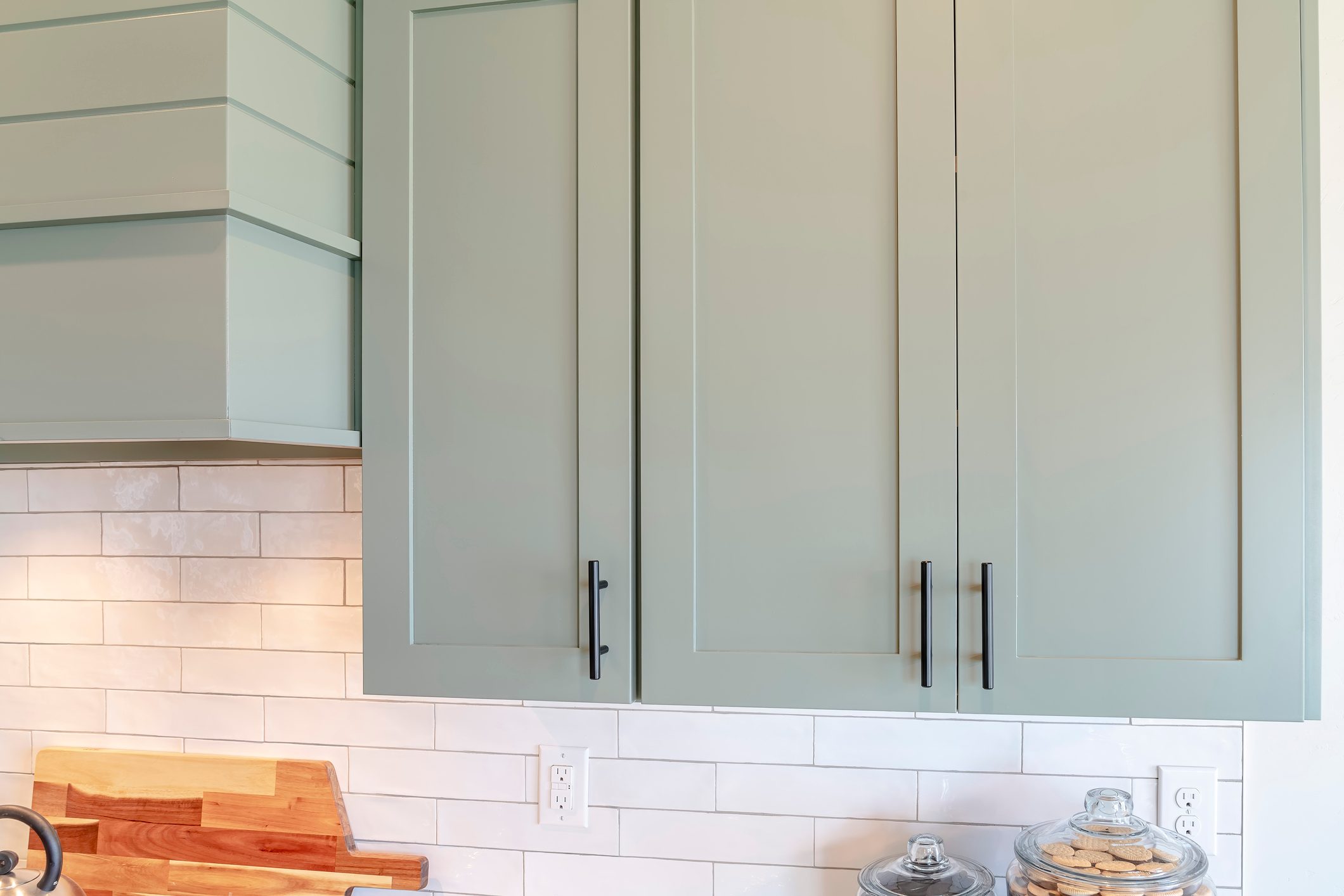Green kitchen cabinets with black handles store items, above a light tiled backsplash, next to a wooden cutting board and glass jars filled with cookies.