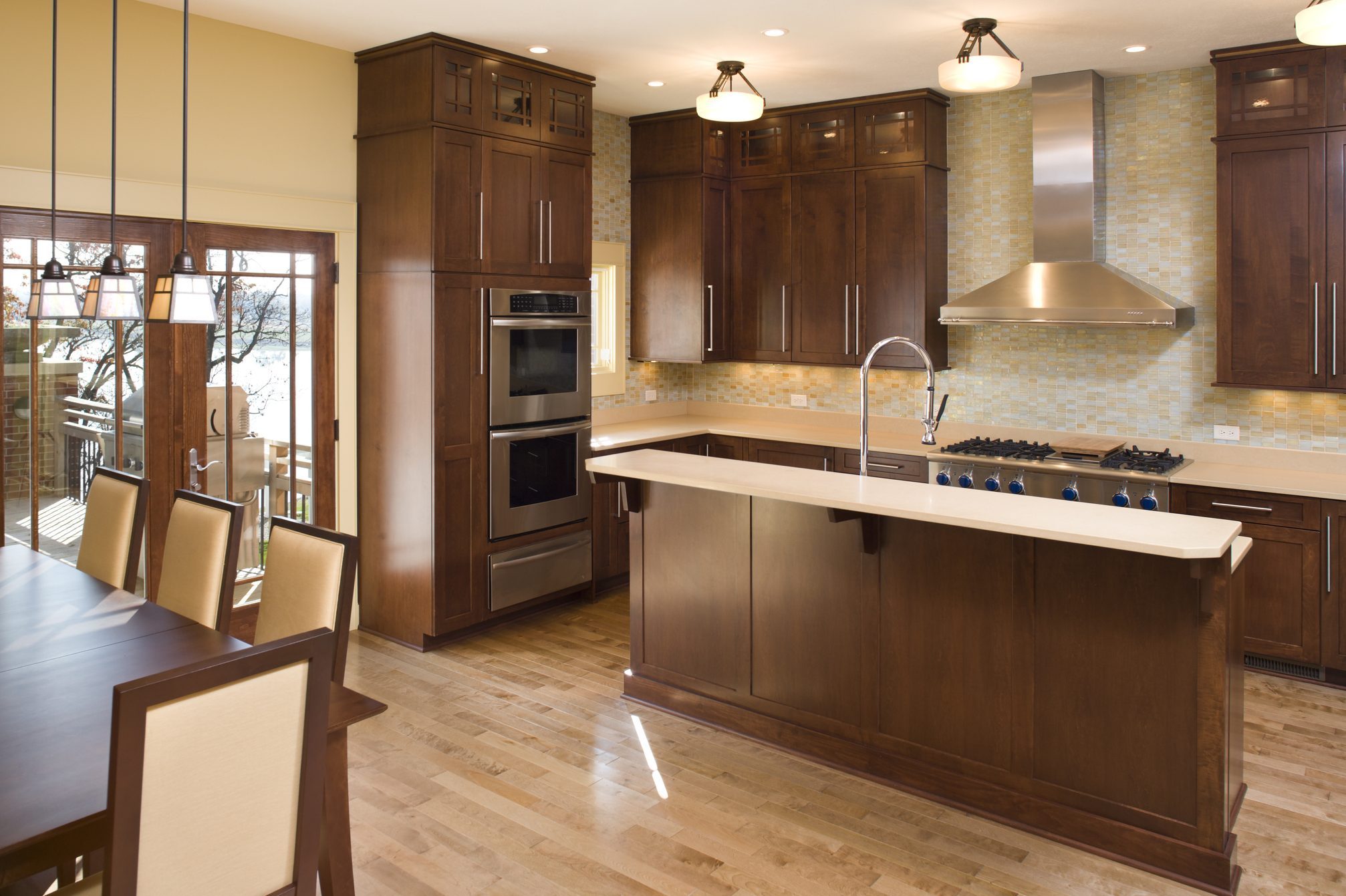A modern kitchen features wooden cabinets, stainless steel appliances, and a large island, illuminated by pendant lights, with a dining area and outdoor view.