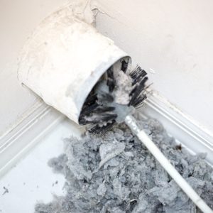 A lint brush is partially inserted into a dusty wall vent, with gray lint and debris accumulating on the floor nearby.