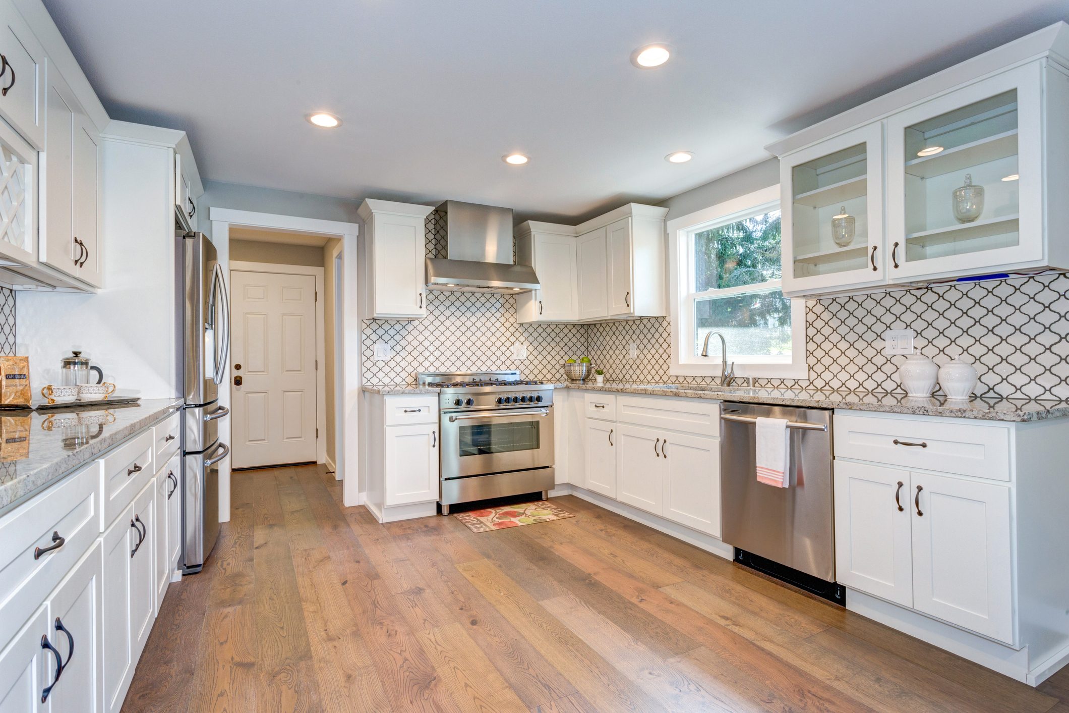 A modern kitchen features stainless steel appliances, white cabinets, and wood flooring, with natural light streaming through the window and a door leading outside.