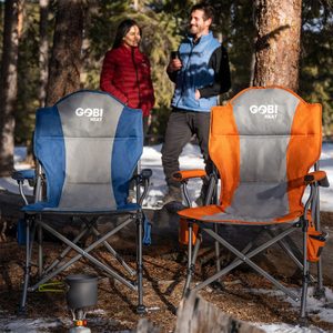 Two folding chairs, one blue and one orange, are placed on a snowy forest floor while two people chat nearby, enjoying the outdoors.