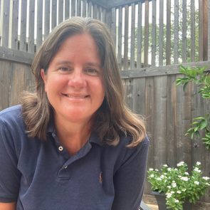 A woman with long hair smiles, sitting outdoors near a wooden fence, surrounded by potted flowers and greenery in a bright, natural setting.