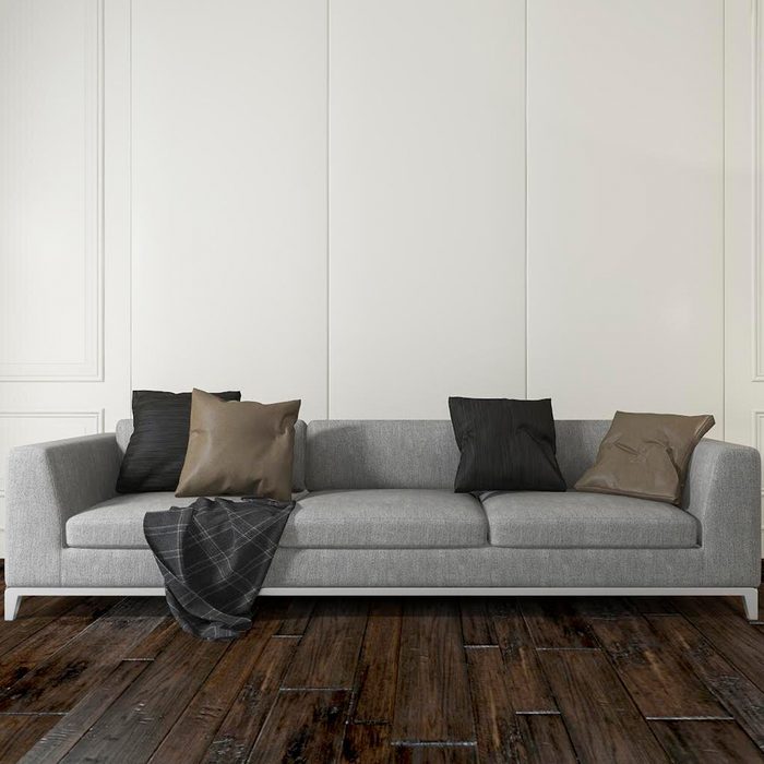 A gray sofa with pillows rests on a wooden floor, accompanied by a blanket draped casually over one side, against a light-colored wall.