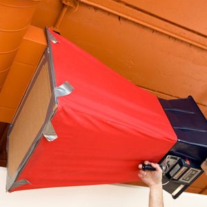 A person holds a black object wrapped in red material, possibly modifying it, under an orange ceiling with pipes above.