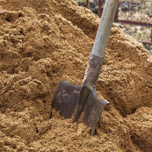 A shovel is embedded in a large pile of brown, granular material, indicating a task of digging or moving soil in a natural outdoor setting.