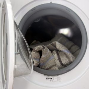 A washing machine door is open, revealing a crumpled towel inside the drum, amidst a blurred background of the laundry room.