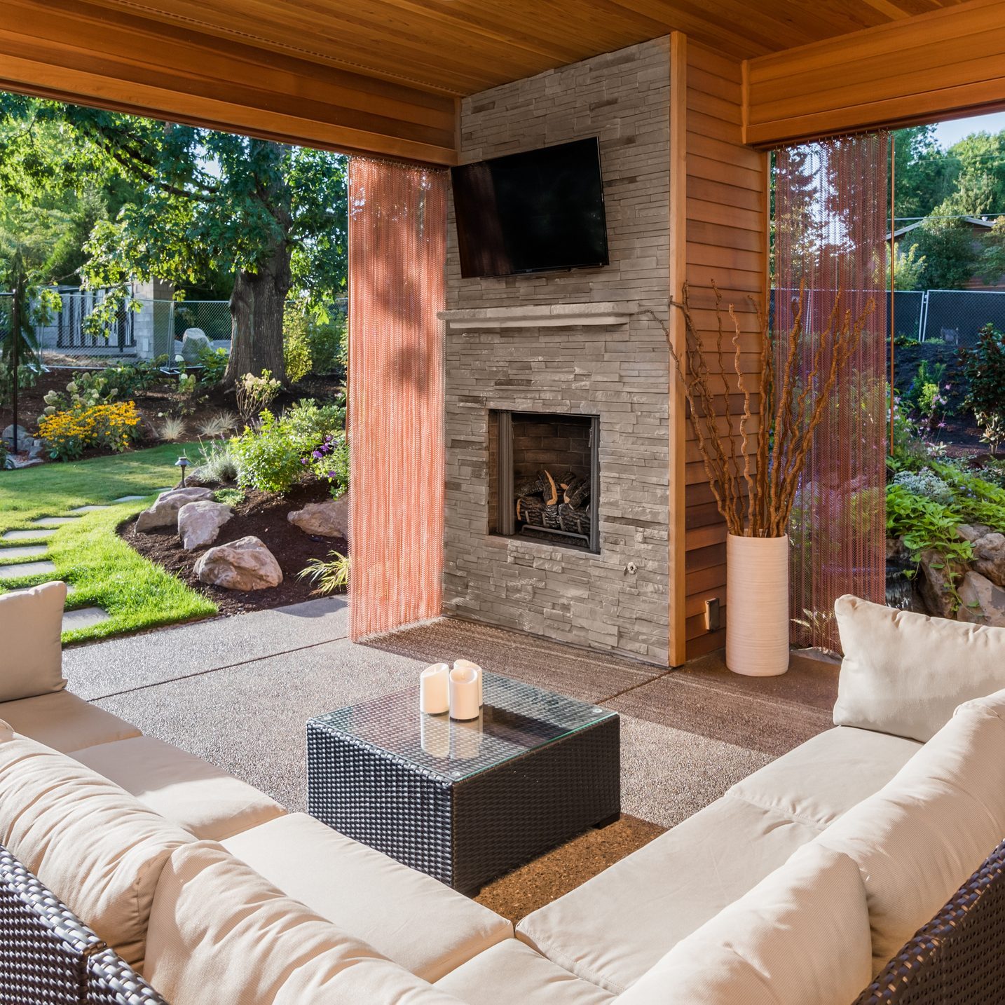 A modern outdoor seating area features a stone fireplace, candles on a glass table, and a view of a garden through sheer curtains.