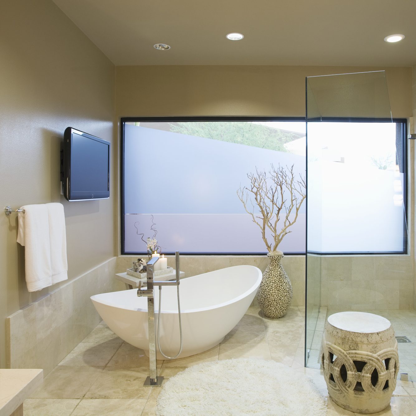 A modern bathroom features a freestanding white tub, chrome faucet, and towel rack, with a decorative vase, a small stool, and large window providing natural light.