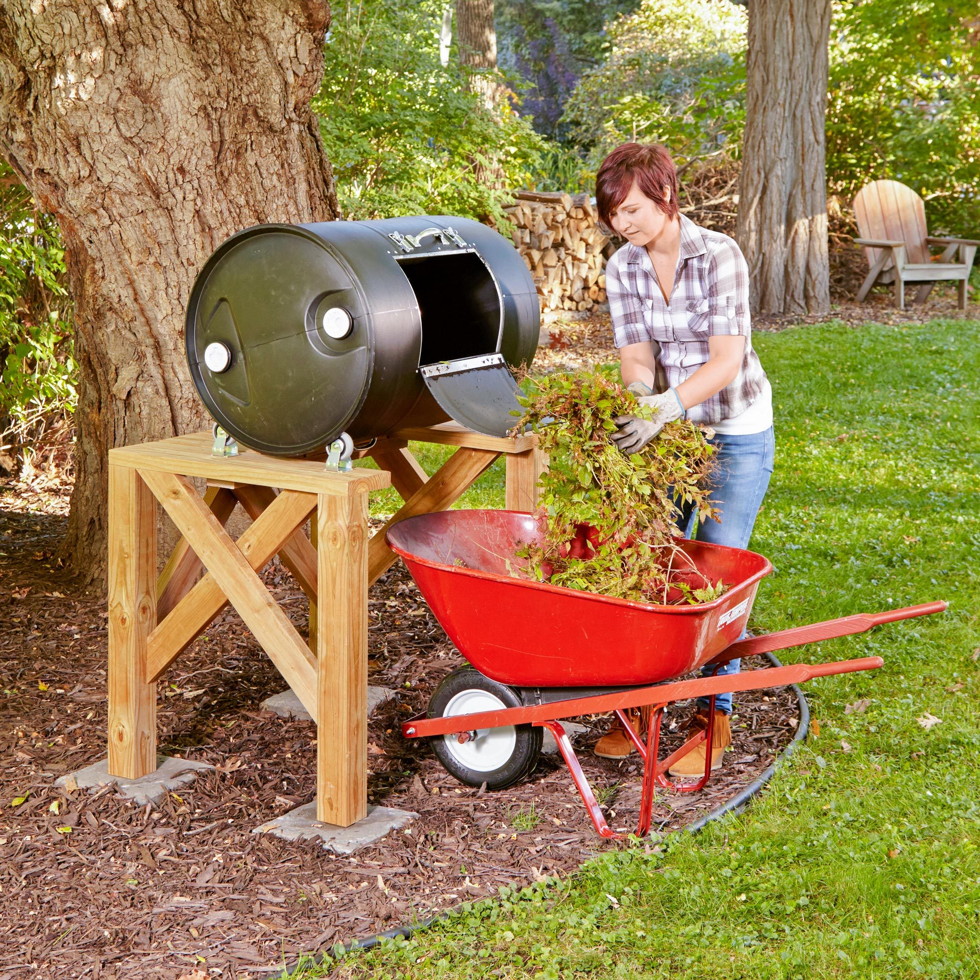 A person discards plant waste into a black compost barrel supported by wooden legs, while a red wheelbarrow sits nearby on grass.