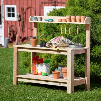 A wooden potting table displays gardening tools, pots, soil bags, and flowers outdoors, surrounded by green grass and a red barn in the background.