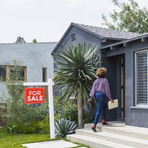A woman carrying documents walks up steps towards a gray house, passing a 