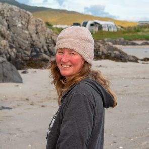 A woman with long, wavy hair, wearing a light-colored beanie and dark hoodie, smiles while standing on a beach near rocky formations and distant trailers.