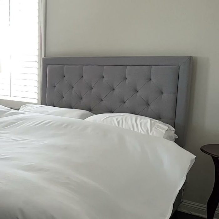 A gray tufted headboard supports white bedding on a bed, illuminated by natural light from a nearby window, with a bedside table visible.
