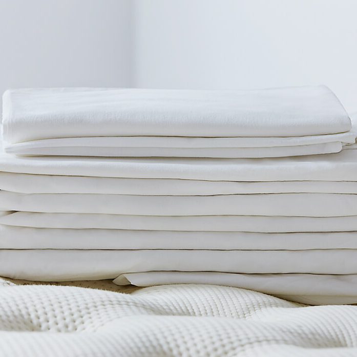A stack of neatly folded white sheets rests on a textured cream-colored blanket, against a softly lit, minimalistic background.
