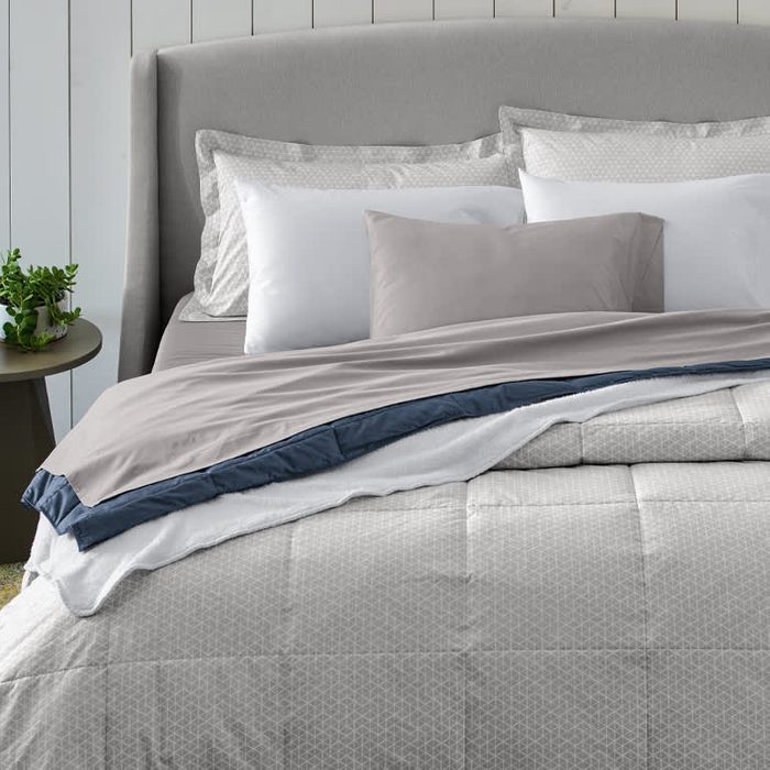 A neatly made bed features layered linens, including a light gray comforter with a dark navy trim, surrounded by decorative pillows against a light wall.
