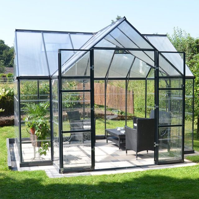 A glass greenhouse stands in a garden, featuring a table and chairs inside. Lush greenery surrounds the structure, creating a serene outdoor space.