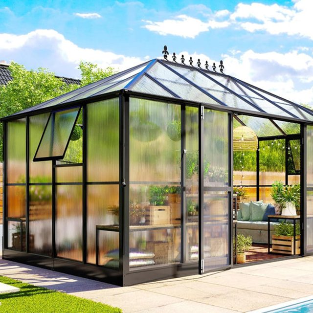 A greenhouse stands with glass walls and a slanted roof, filled with plants inside, surrounded by a landscaped garden and blue sky.