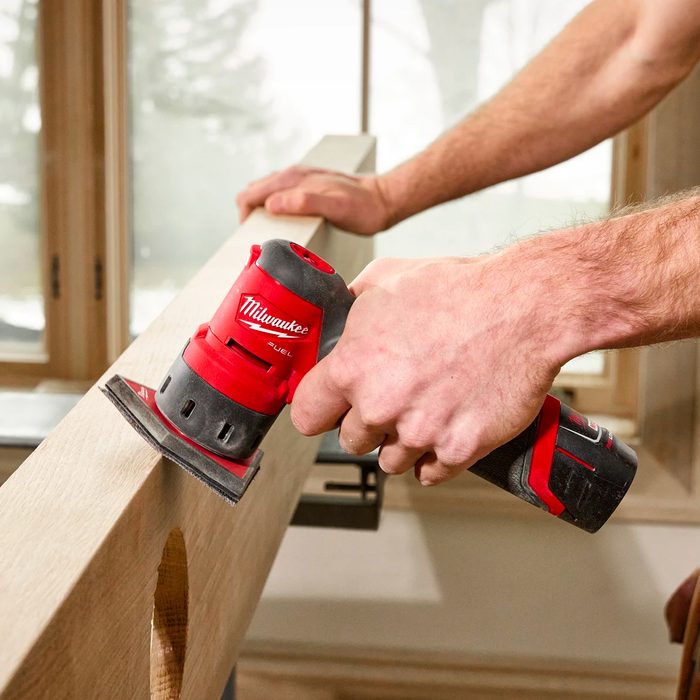 A person uses a red and black power sander on a wooden board, refining its surface in a well-lit room with large windows.