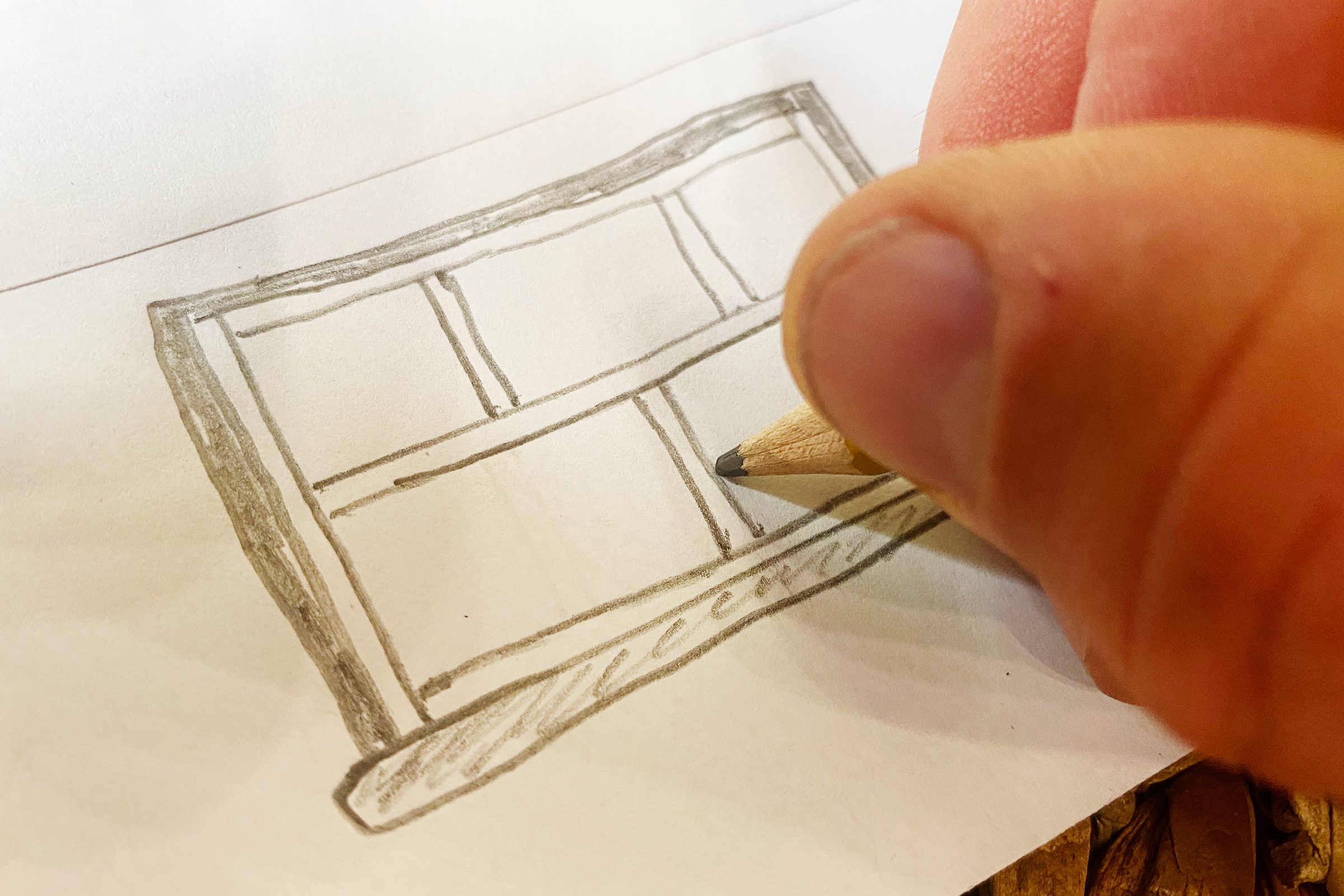 A hand holds a pencil, sketching a rectangular window on white paper, surrounded by a textured surface.