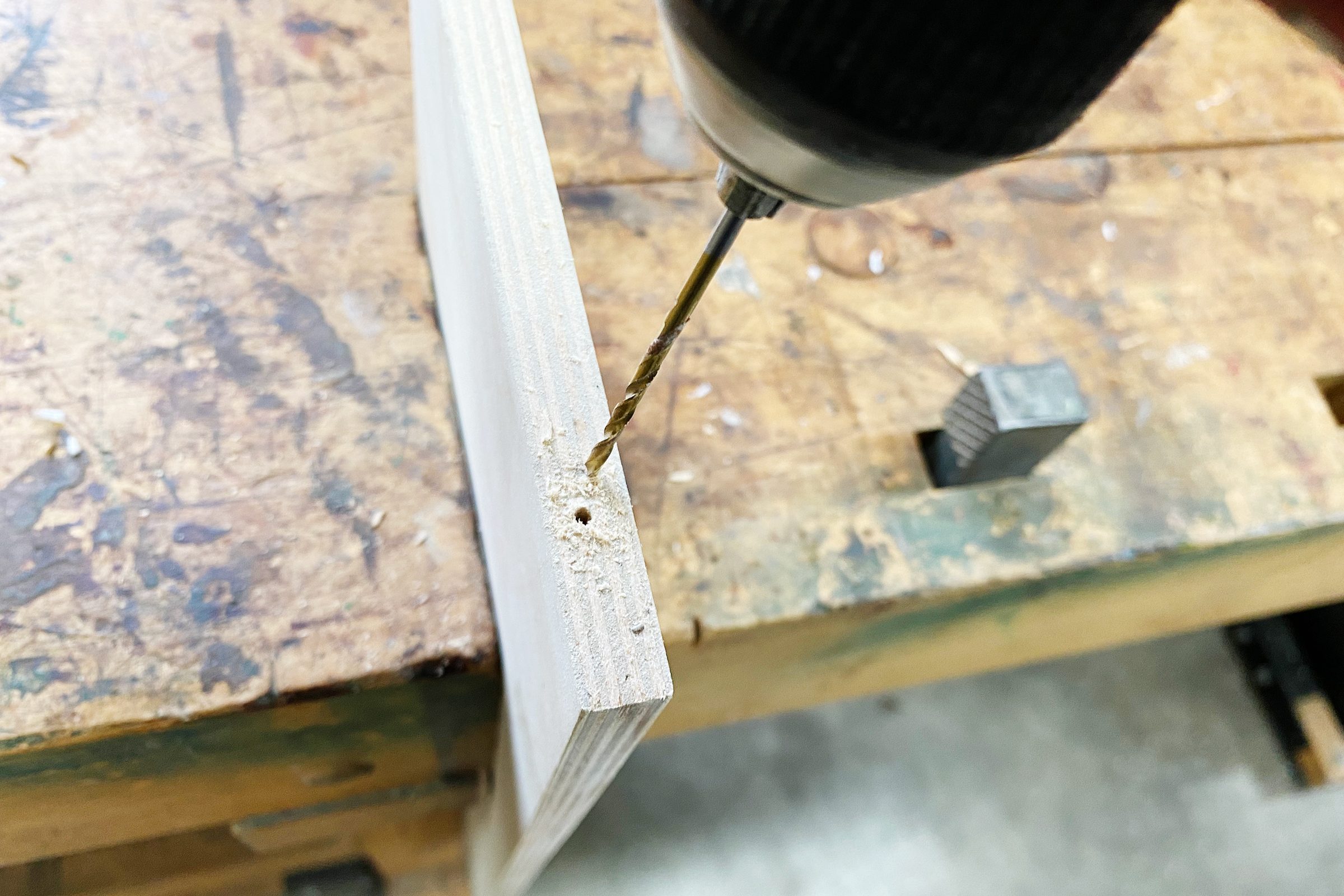 A drill is creating a hole in the edge of a piece of wood, resting on a workbench with visible wear marks.