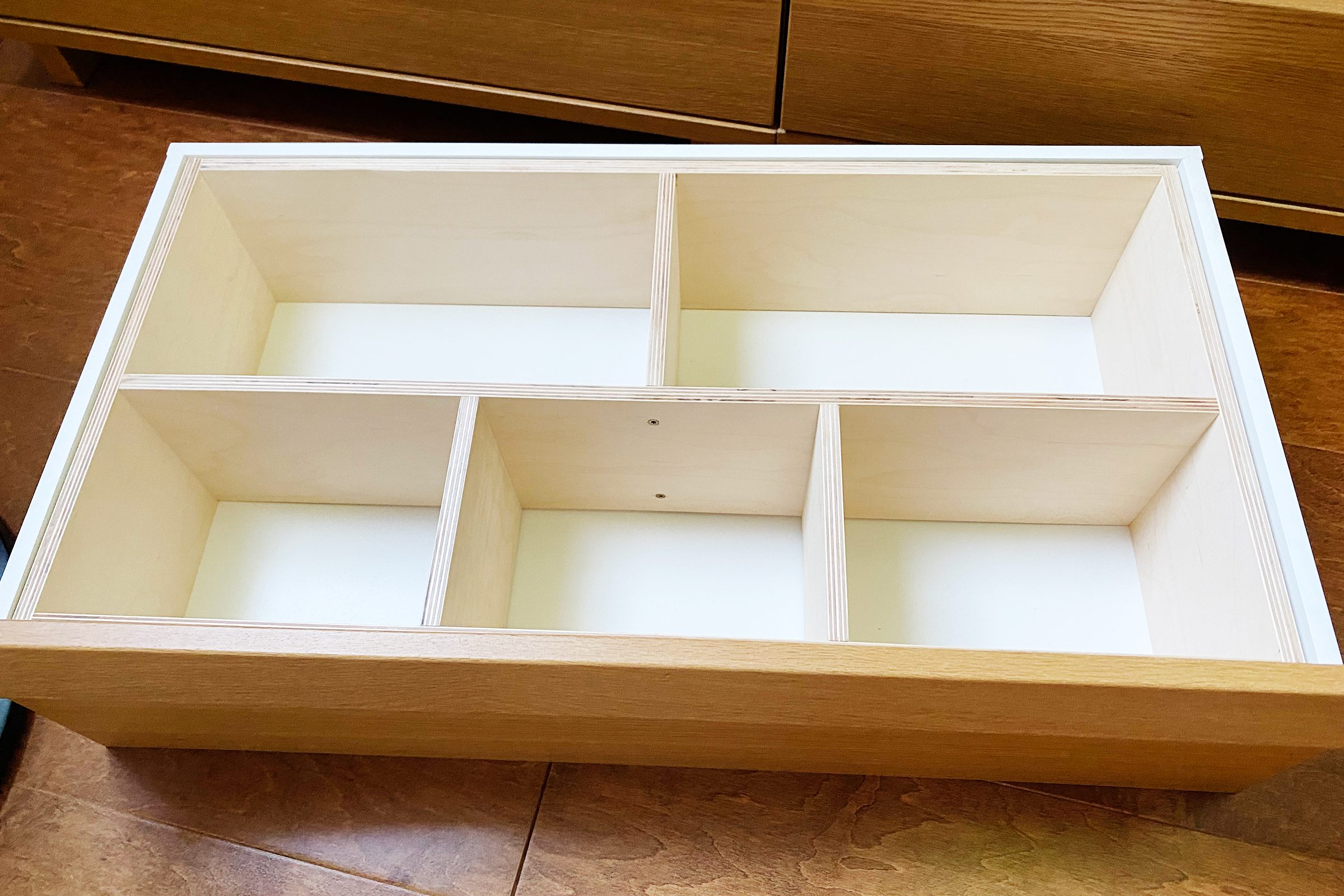 A wooden drawer organizer with six compartments sits on a brown wooden floor, ready for storage. The compartments are empty and neatly outlined.