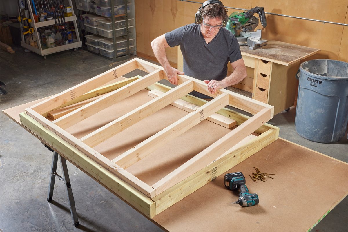 A person is assembling wooden frames on a workbench in a workshop, with tools and materials around, including a drill and nails.