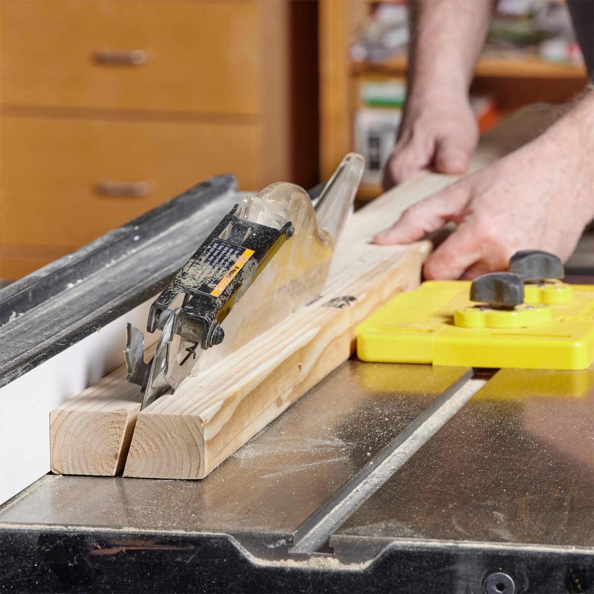 A saw is cutting into a wooden plank, while a person stabilizes it on a table saw in a workshop filled with tools and cabinetry.