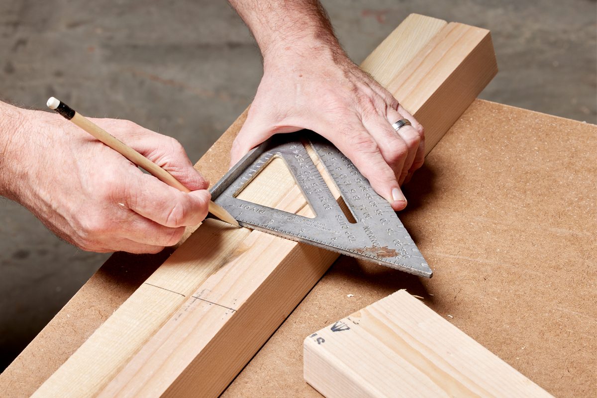 A carpenter's hand holds a square while marking a line on wood, positioned on a workbench in a workshop environment.