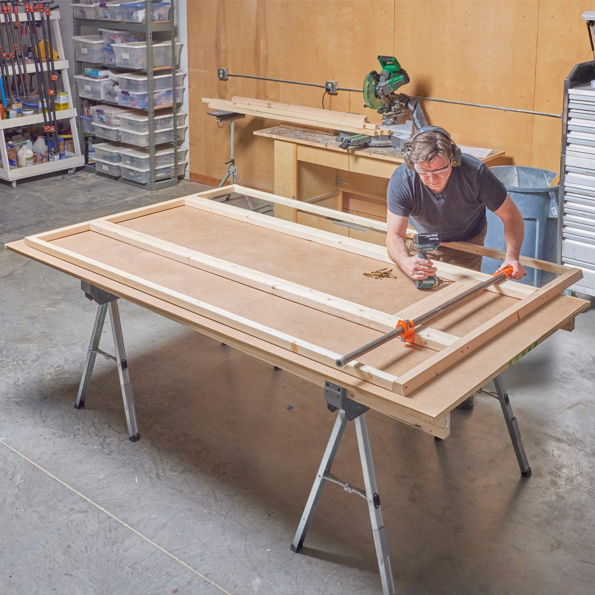 A person uses a power drill to secure wood pieces on a workbench in a workshop, surrounded by tools and storage containers.