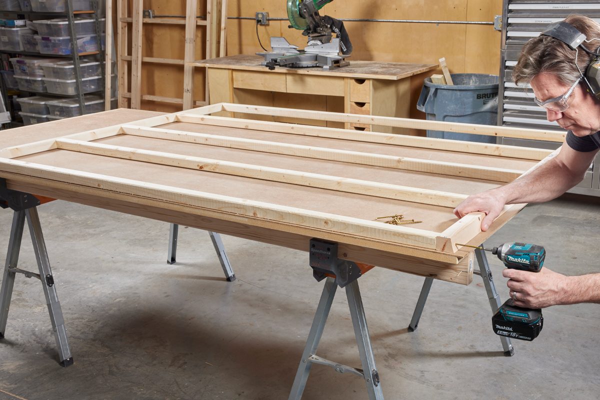 A man screws wooden beams into a frame on a worktable in a workshop, surrounded by storage bins and tools.