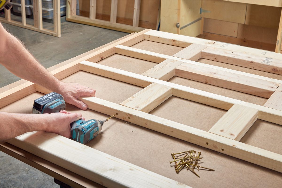 A hand uses a power drill to secure wooden beams on a flat surface, surrounded by additional wooden pieces and scattered screws in a workshop environment.