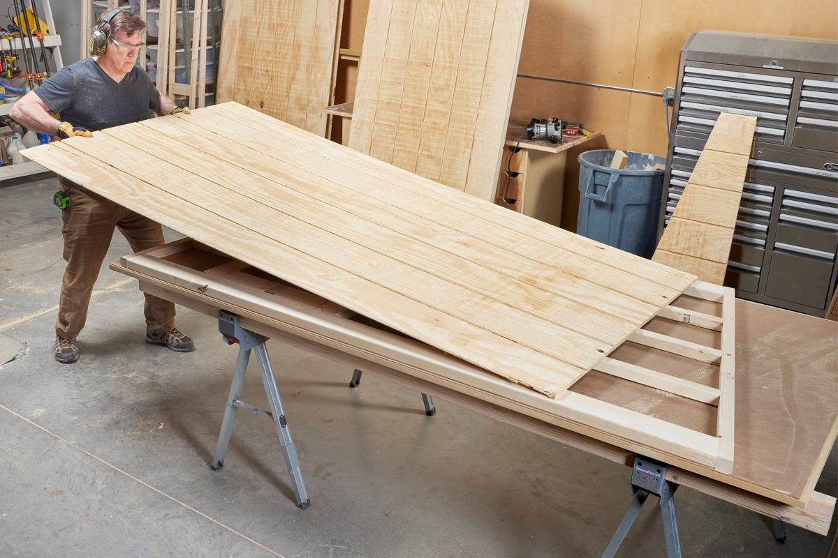 A worker lifts a large wooden panel above a sawhorse in a workshop, surrounded by tools and wood materials.