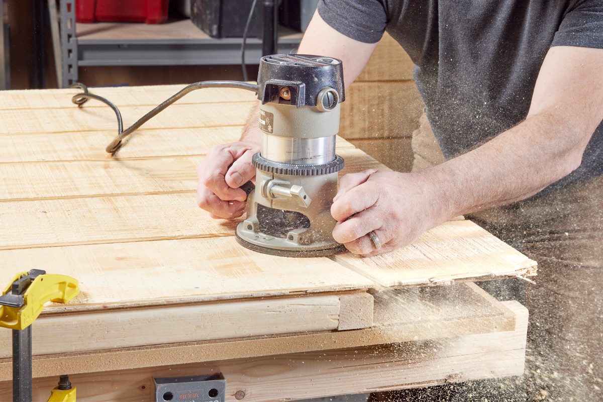 A router is used to shape wood planks on a workbench, creating sawdust in a well-lit workshop environment.
