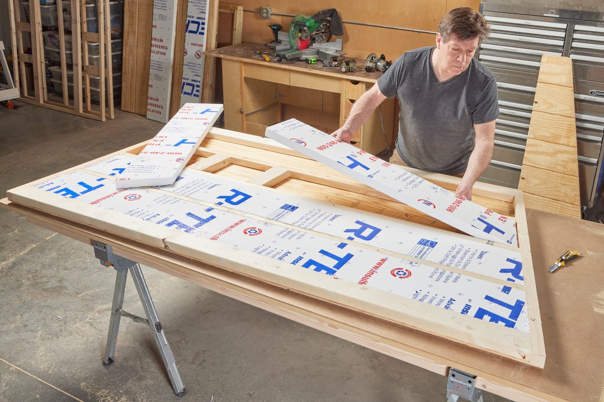 A man arranges insulation sheets on a wood frame in a workshop, surrounded by tools and wooden materials on work surfaces.