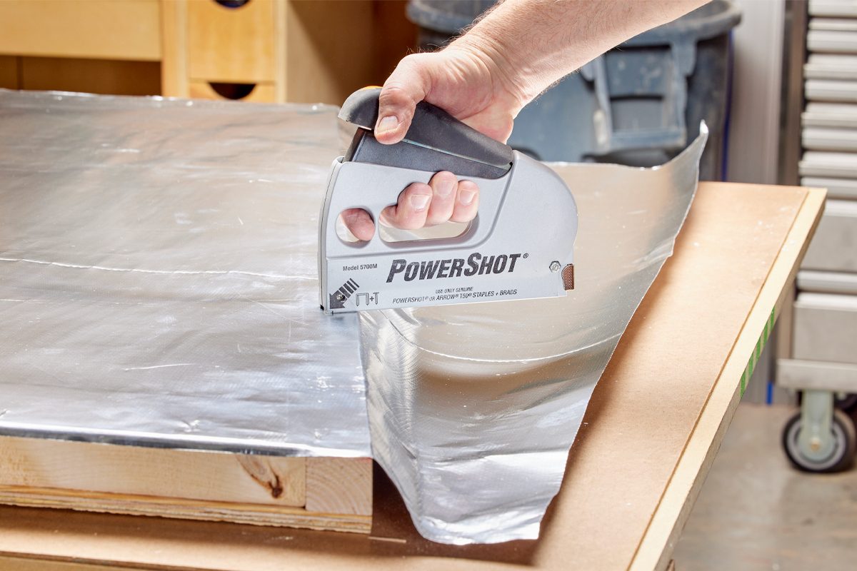 A hand holds a staple gun, applying it to aluminum sheeting on a wooden surface in a workshop environment. Tools and storage units are visible in the background.