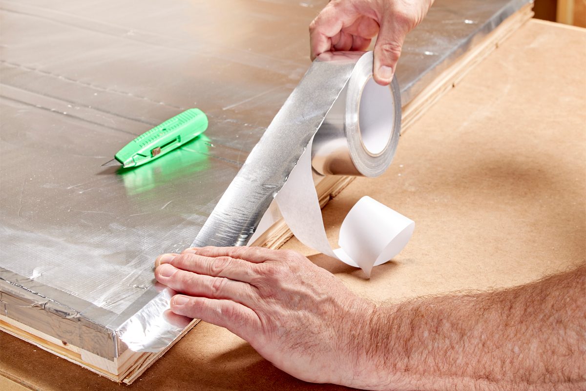 A hand applies aluminum tape to an edge of a silver surface while a utility knife rests nearby on a wooden table.