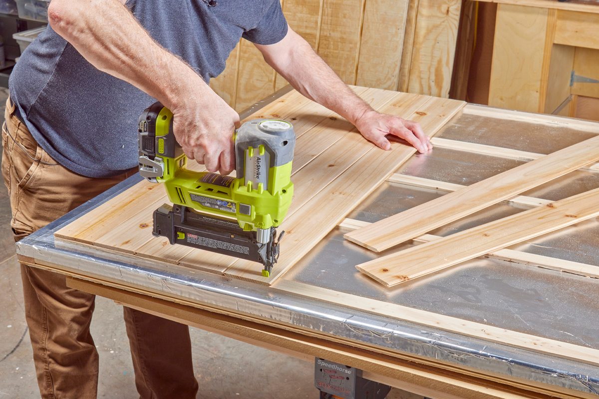 A person is using a power nailer to fasten wooden planks on a work table in a workshop, surrounded by wooden materials and tools.